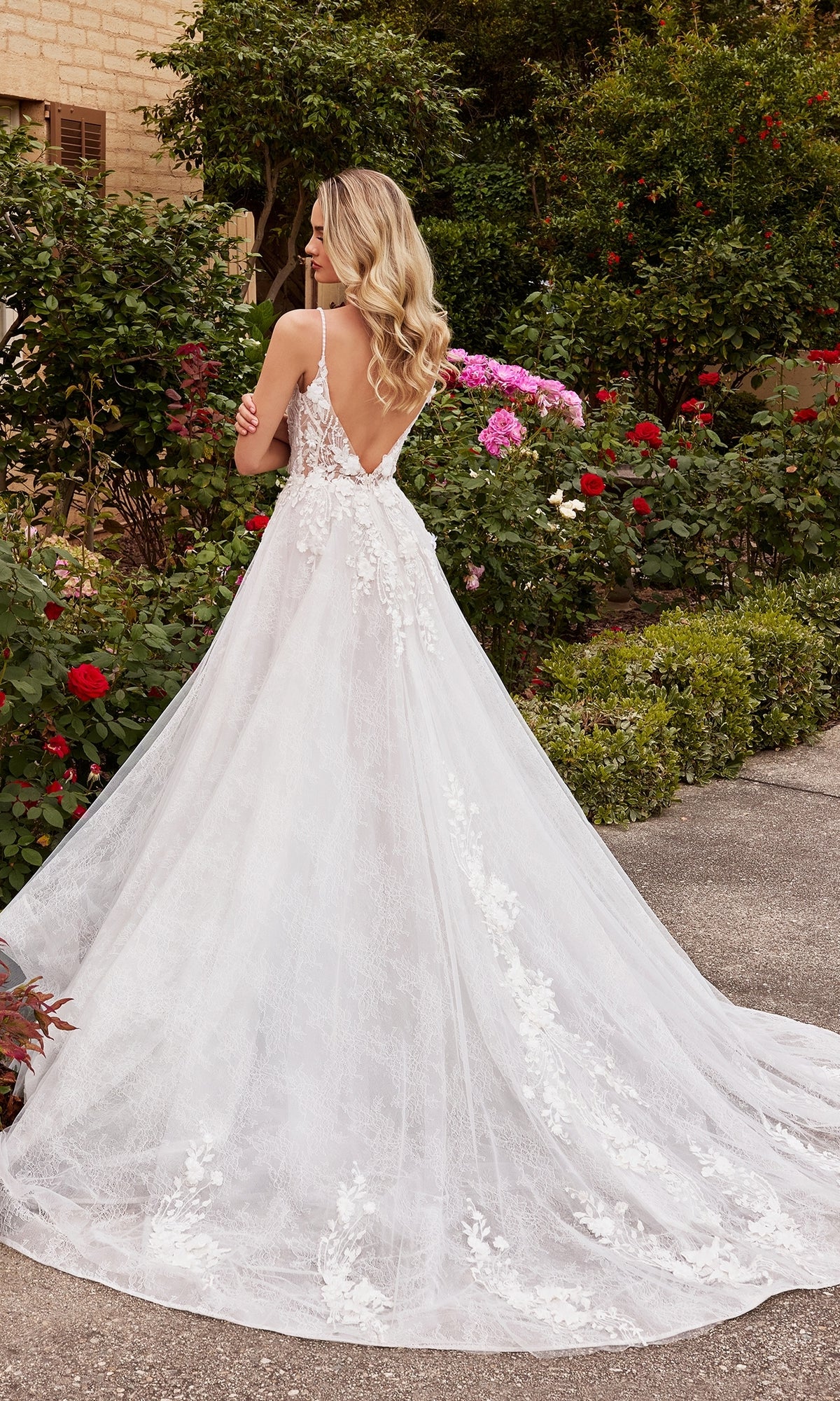 A woman wearing the A-Line White Lace Wedding Gown CD857W with a sheer corset bodice stands outdoors in a garden, surrounded by blooming roses and lush greenery.